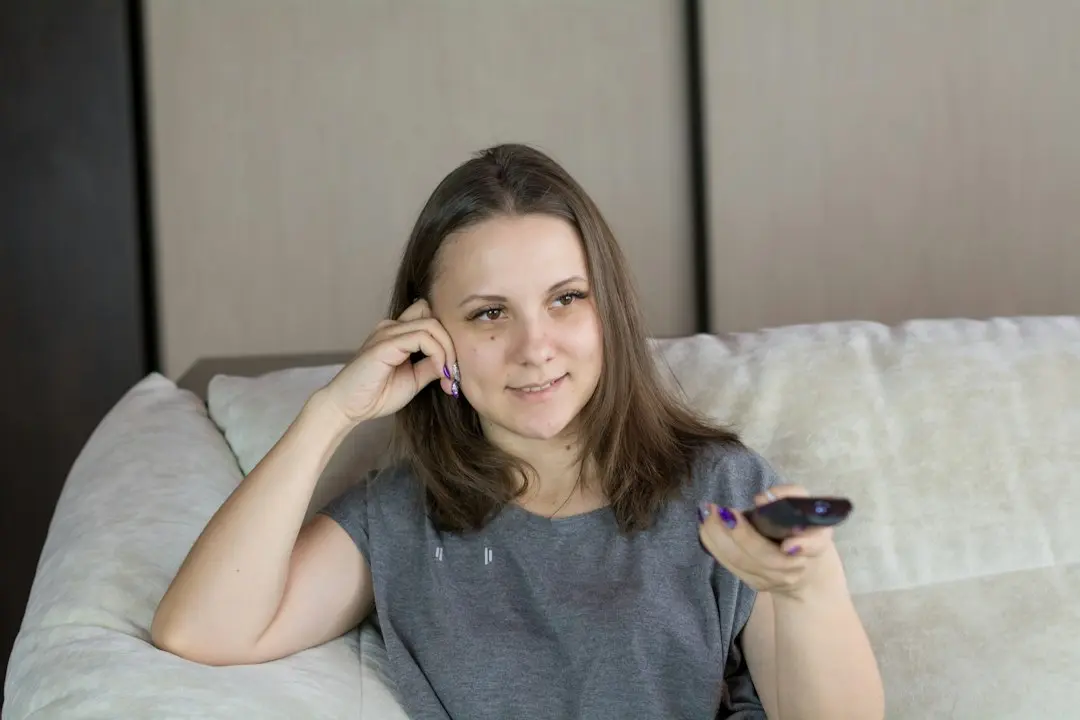 A smiling woman on a light sofa pointing a remote at the TV, as when judging which IPTV service suits the household