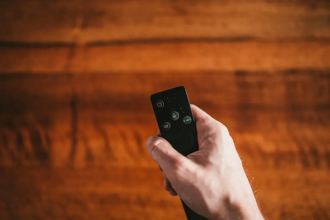 A hand holding a black TV remote against a warm wooden wall, as when browsing IPTV channels